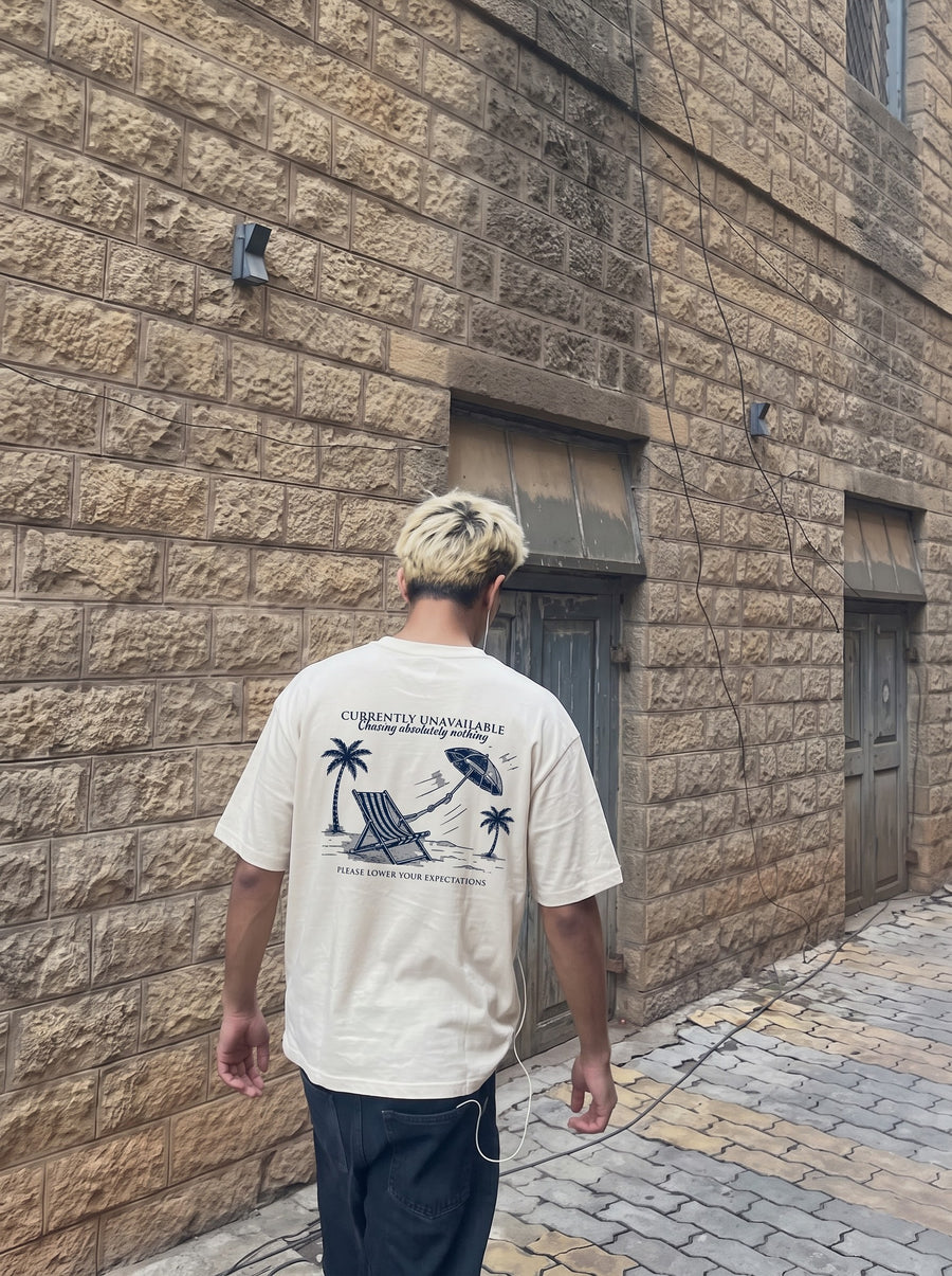Currently Unavailable back print on an off-white t-shirt, worn on a cobbled street beside a brick wall