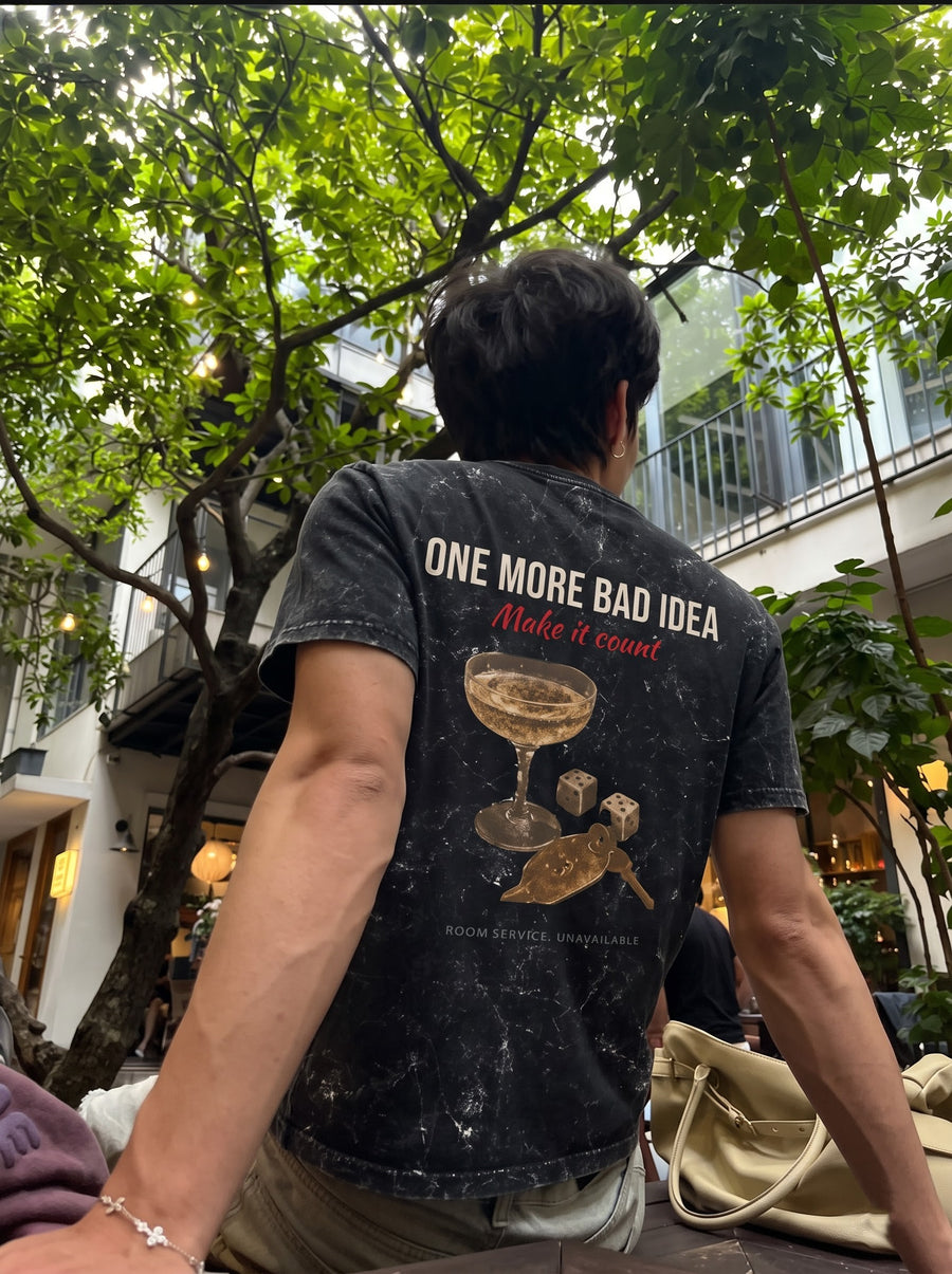 One More Bad Idea back print with a coupe glass and dice, on a black acid-wash t-shirt, worn under trees