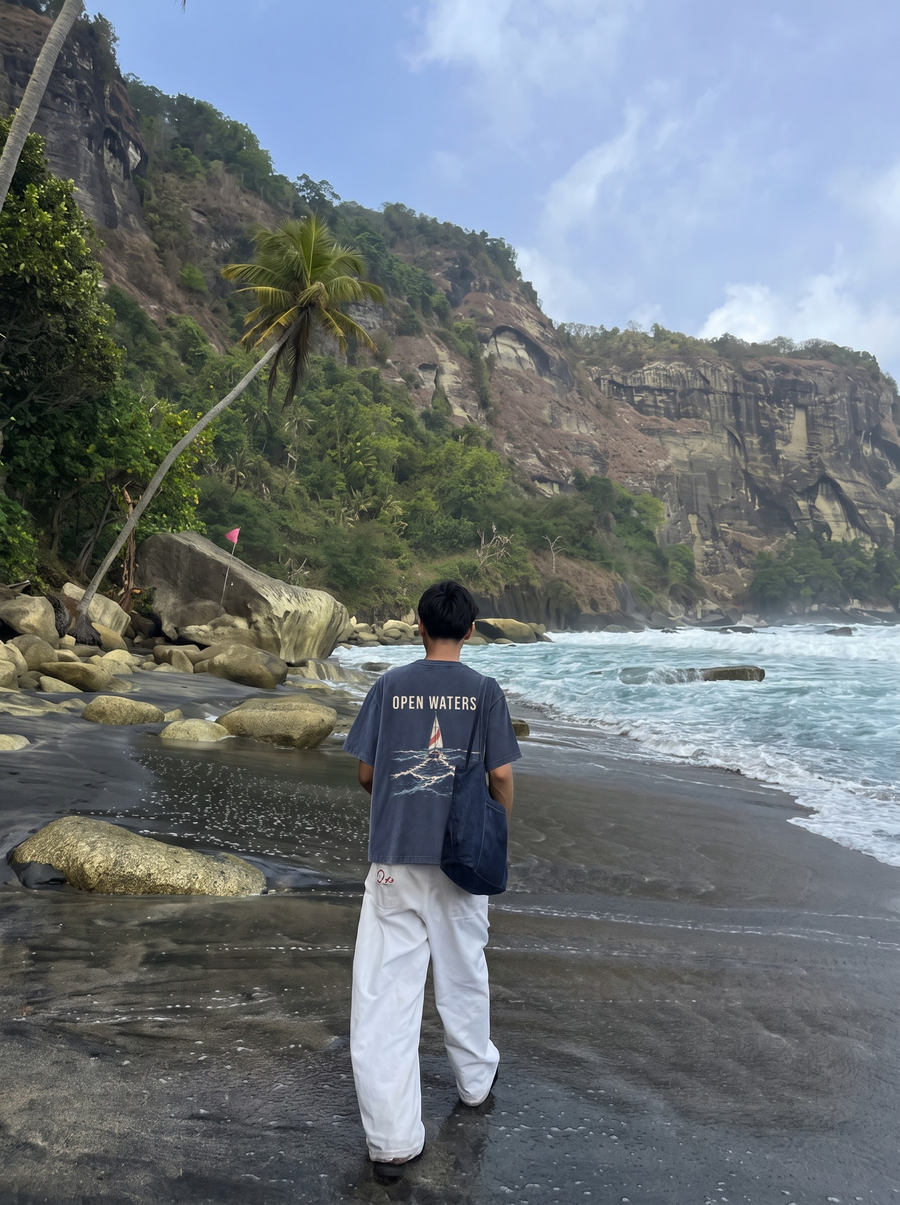 Open Waters back print on a navy acid-wash t-shirt, worn on a beach below cliffs