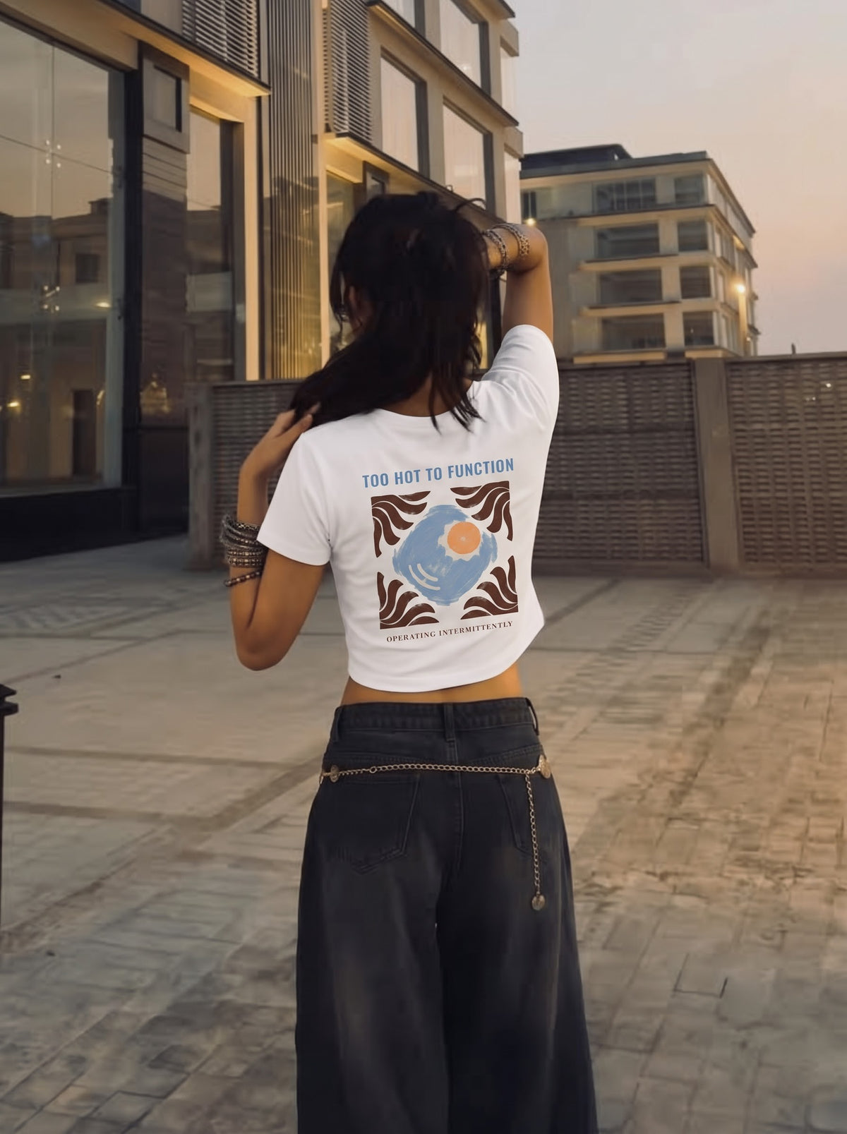 Too Hot To Function baby tee in white, worn from behind with dark wide-leg jeans at dusk, the sun-tile back print and the words operating intermittently