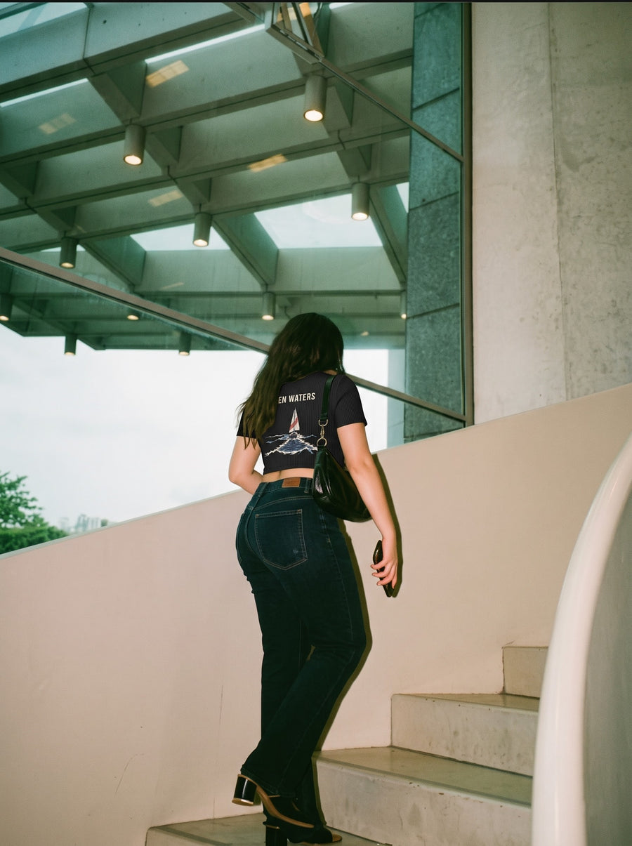 Open Waters baby tee in black, worn from behind climbing a staircase in dark jeans, the sailboat back print showing
