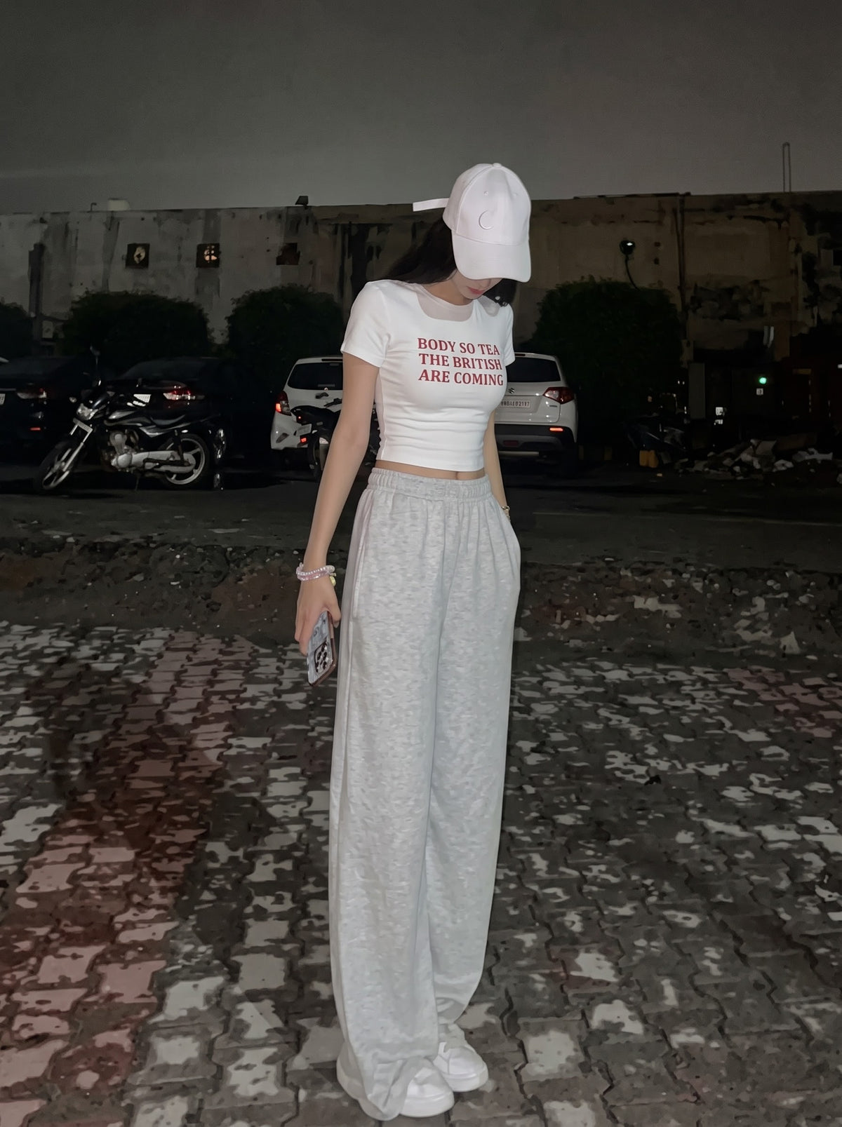 British Are Coming baby tee in white, worn with grey wide-leg joggers and a white cap at night, with Body so tea, the British are coming in red across the front