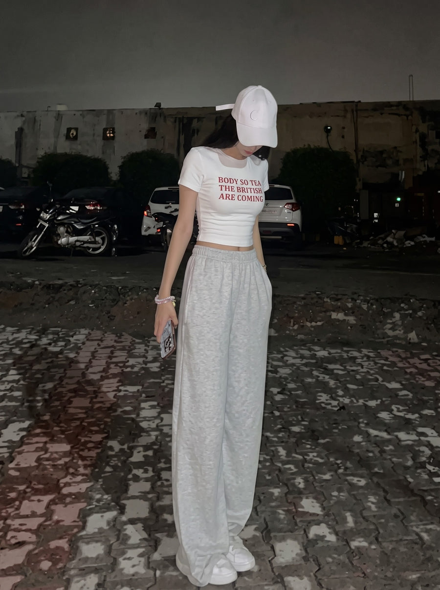 British Are Coming baby tee in white, worn with grey wide-leg joggers and a white cap at night, with Body so tea, the British are coming in red across the front