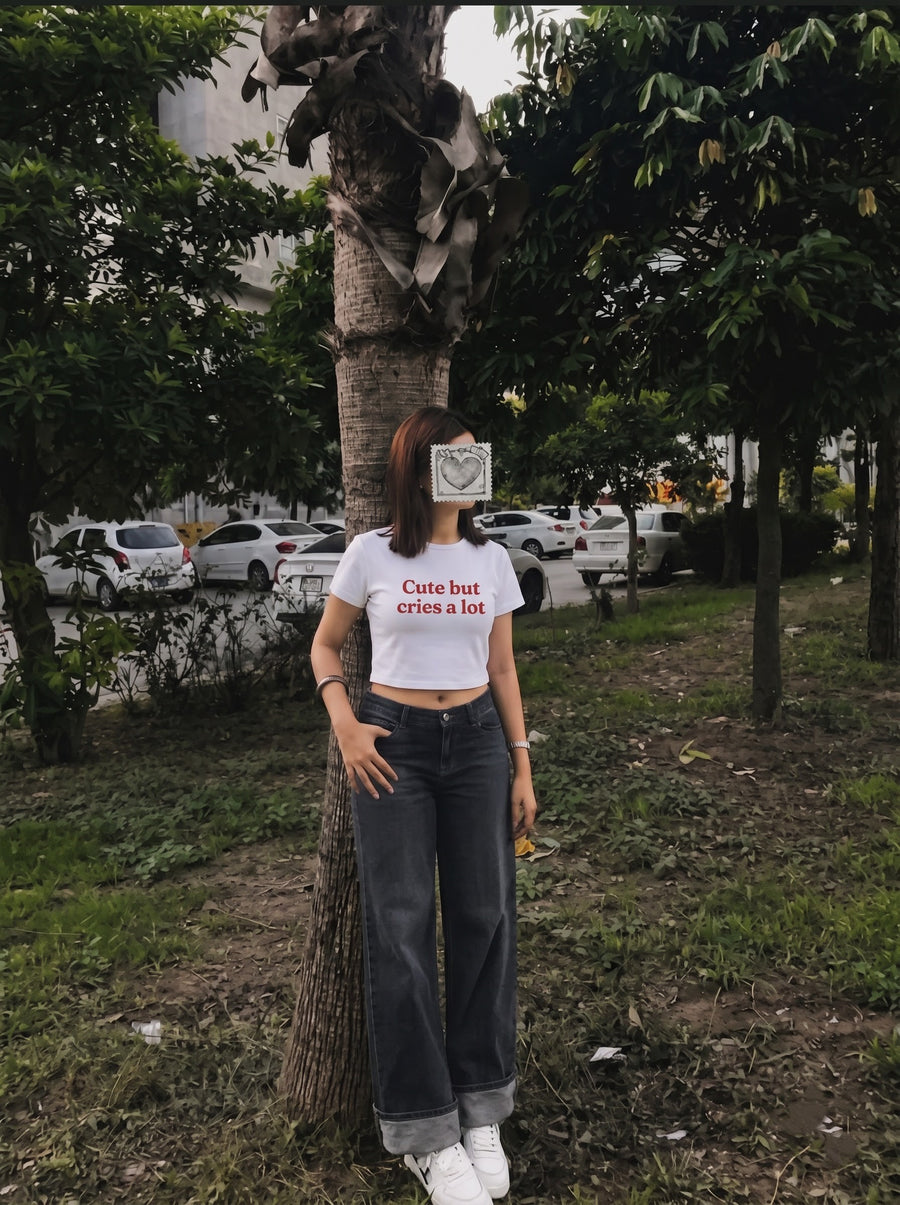 Cute But Cries a Lot baby tee in white, worn with dark wide-leg jeans against a tree in a park, with Cute but cries a lot in red across the front