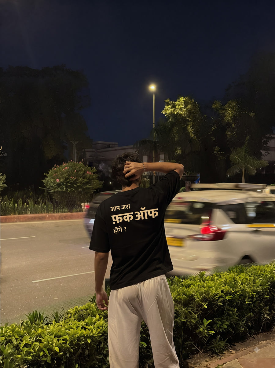 Aap Zara oversized t-shirt in black, worn from behind with white trousers beside a road at night, a street lamp in the distance, three lines of cream Devanagari across the back