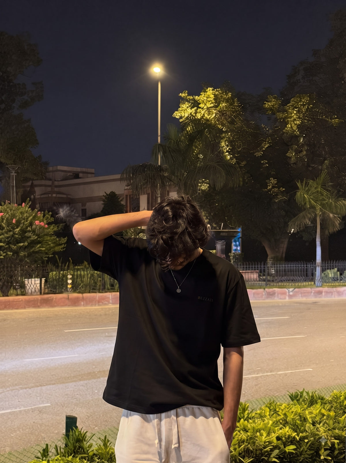 Front of the black Aap Zara oversized t-shirt, worn at night by the roadside with white drawstring trousers and a hand on the head, small BEZZATI wordmark on the left chest