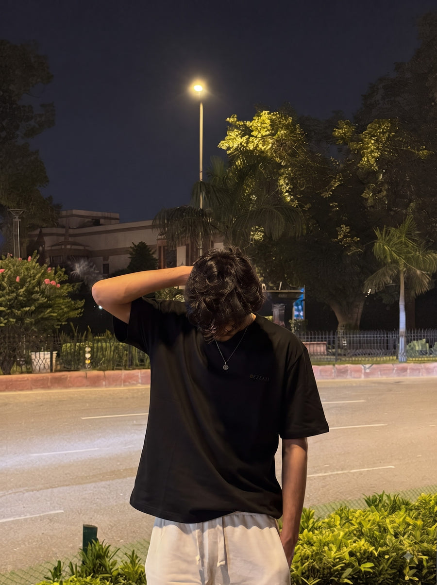 Front of the black Aap Zara oversized t-shirt, worn at night by the roadside with white drawstring trousers and a hand on the head, small BEZZATI wordmark on the left chest