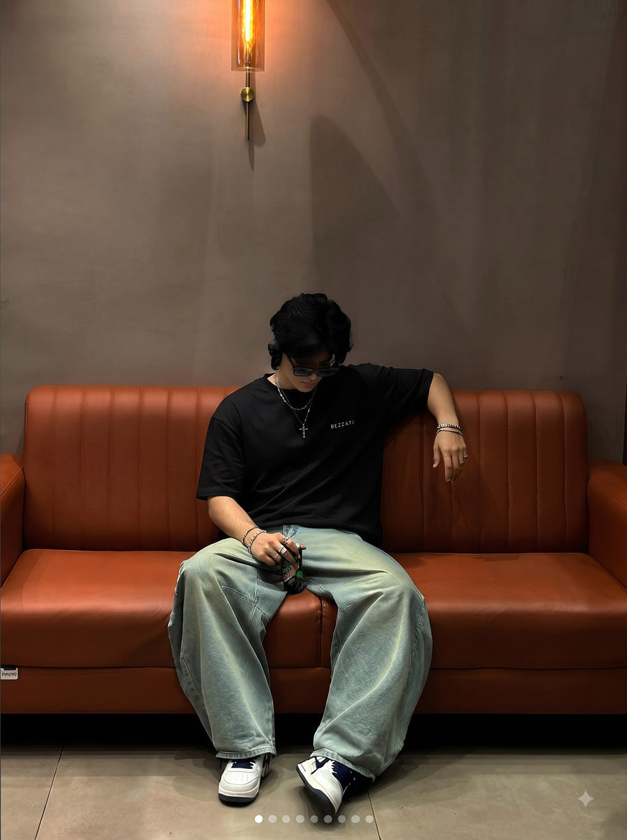 Black Happy Life oversized t-shirt worn seated on a tan leather sofa with wide light-wash jeans, small BEZZATI wordmark on the left chest