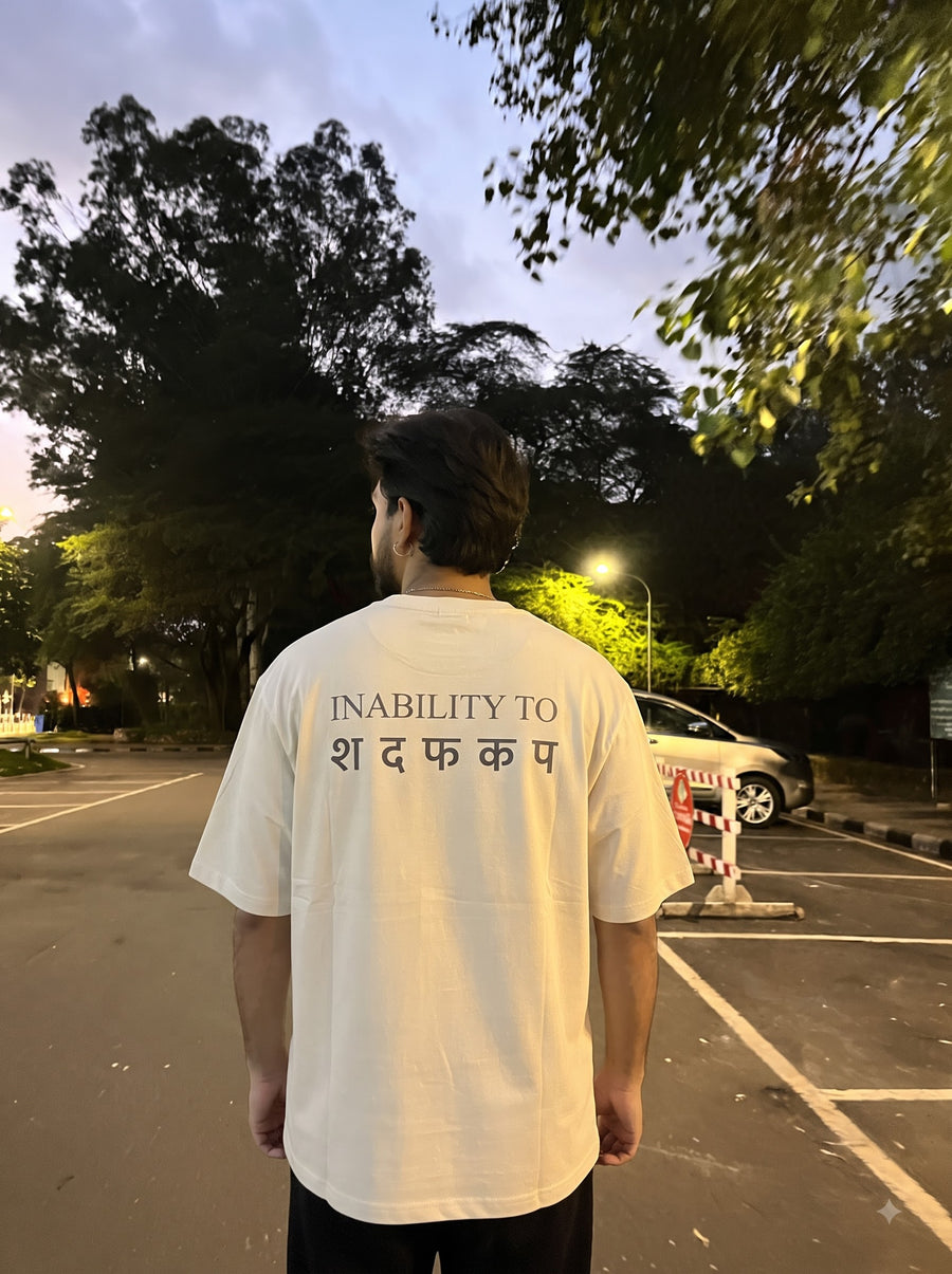 Inability oversized t-shirt in White, worn from behind in a car park at dusk under trees and a street lamp, with INABILITY TO in grey serif capitals above a row of Devanagari letters across the back