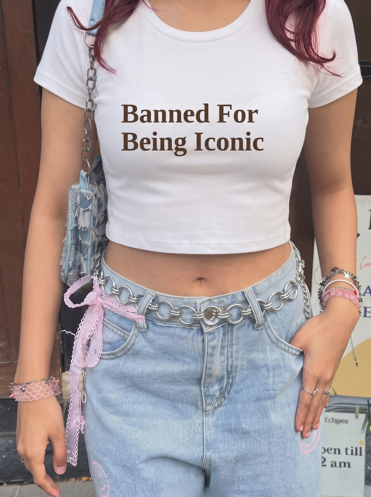 Banned baby tee in White, worn with light-wash jeans, a chain belt tied with a pink lace ribbon and a denim shoulder bag outside a wooden door, with Banned For Being Iconic in dark brown across the front