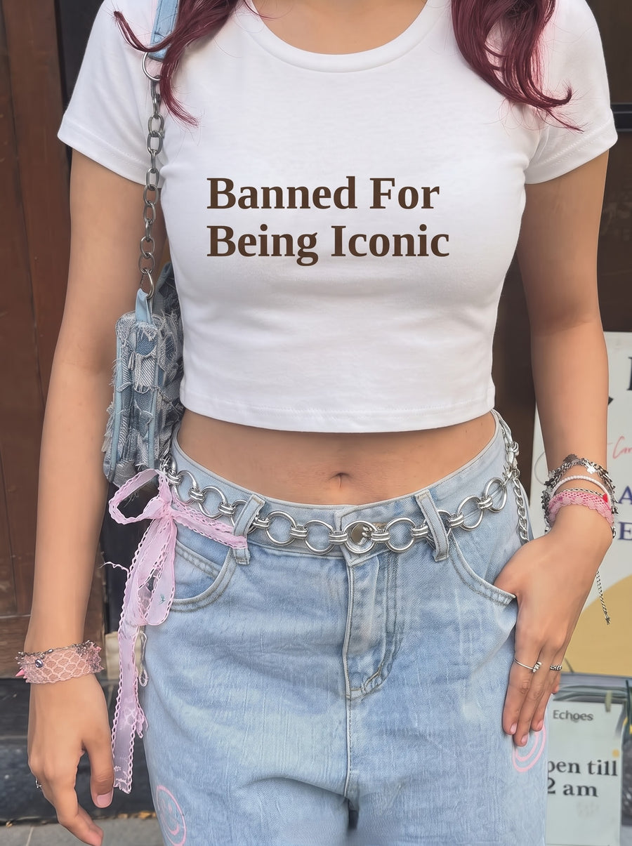 Banned baby tee in White, worn with light-wash jeans, a chain belt tied with a pink lace ribbon and a denim shoulder bag outside a wooden door, with Banned For Being Iconic in dark brown across the front