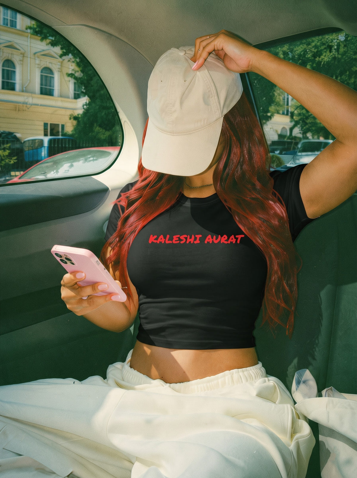 Kaleshi Aurat baby tee in black, worn in the back seat of a car with a beige cap pulled low, long red hair and cream joggers, looking at a pink phone, with Kaleshi aurat in red across the front