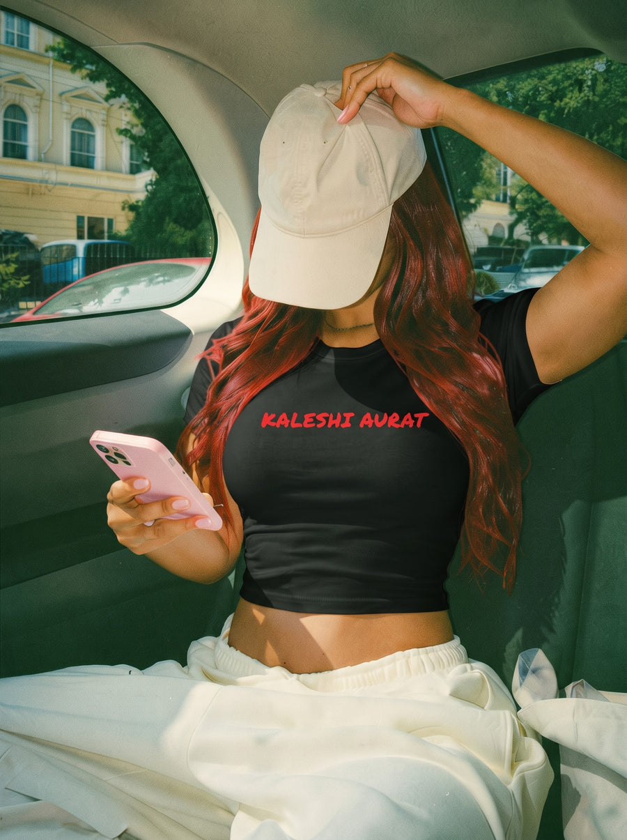 Kaleshi Aurat baby tee in black, worn in the back seat of a car with a beige cap pulled low, long red hair and cream joggers, looking at a pink phone, with Kaleshi aurat in red across the front