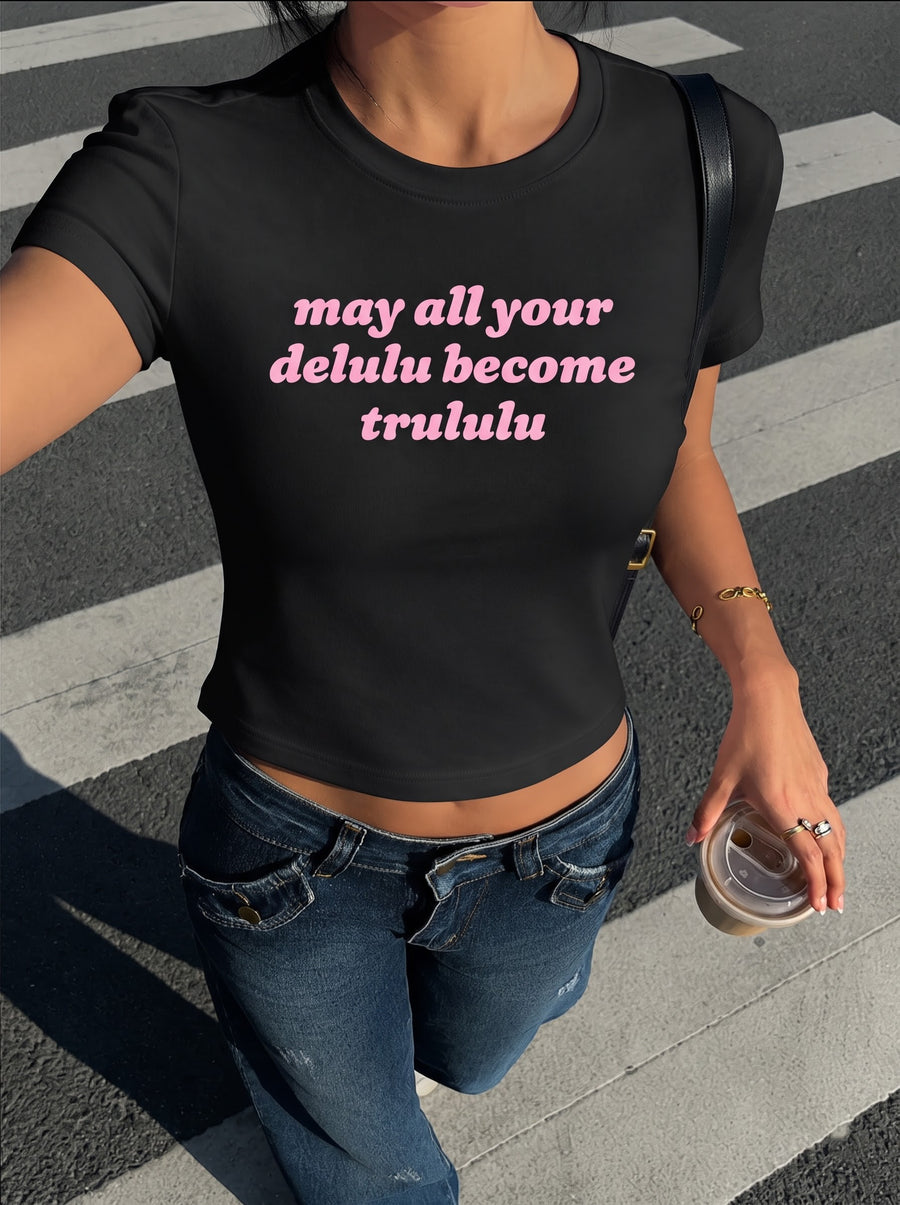 Delulu Become Trululu baby tee in black, worn with low-rise dark jeans and an iced coffee on a zebra crossing, with may all your delulu become trululu in pink across the front