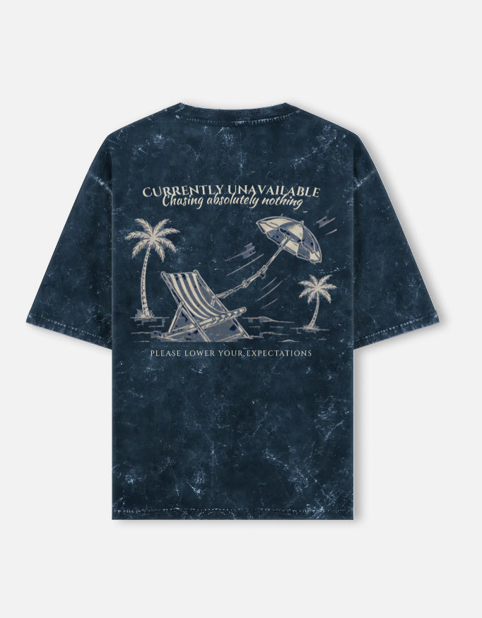 Currently Unavailable back print on a navy acid-wash oversized t-shirt, with a deck chair, palm trees and a beach umbrella blowing away
