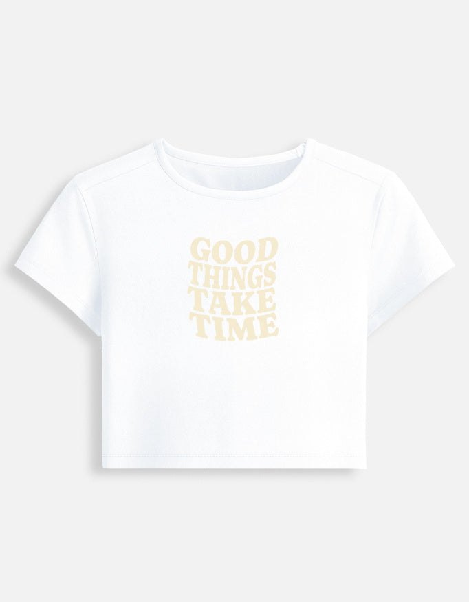 Front of the White Good Things Take Time cropped baby tee: Good things take time in wavy cream serif capitals, a soft, low-contrast print