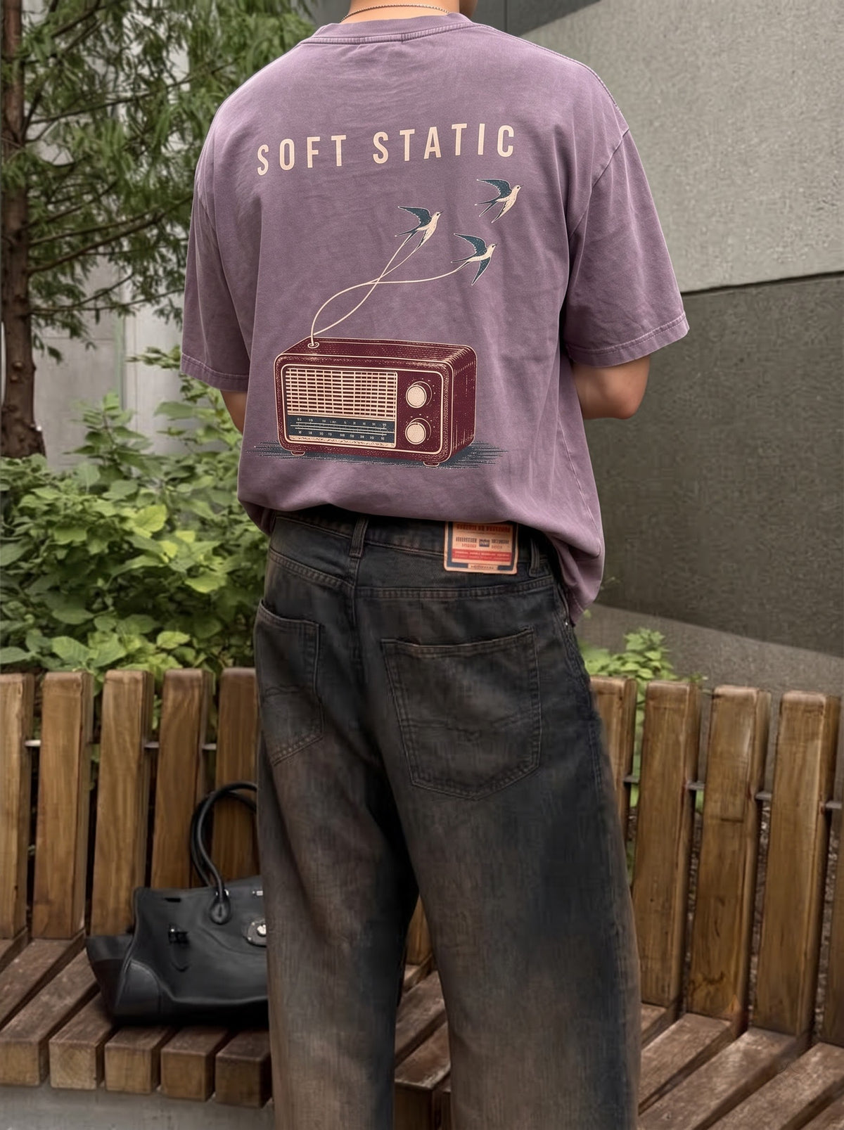 The Soft Static purple oversized tee worn against a wooden fence, an old valve radio with two birds lifting out of the sound printed the full width of the back.