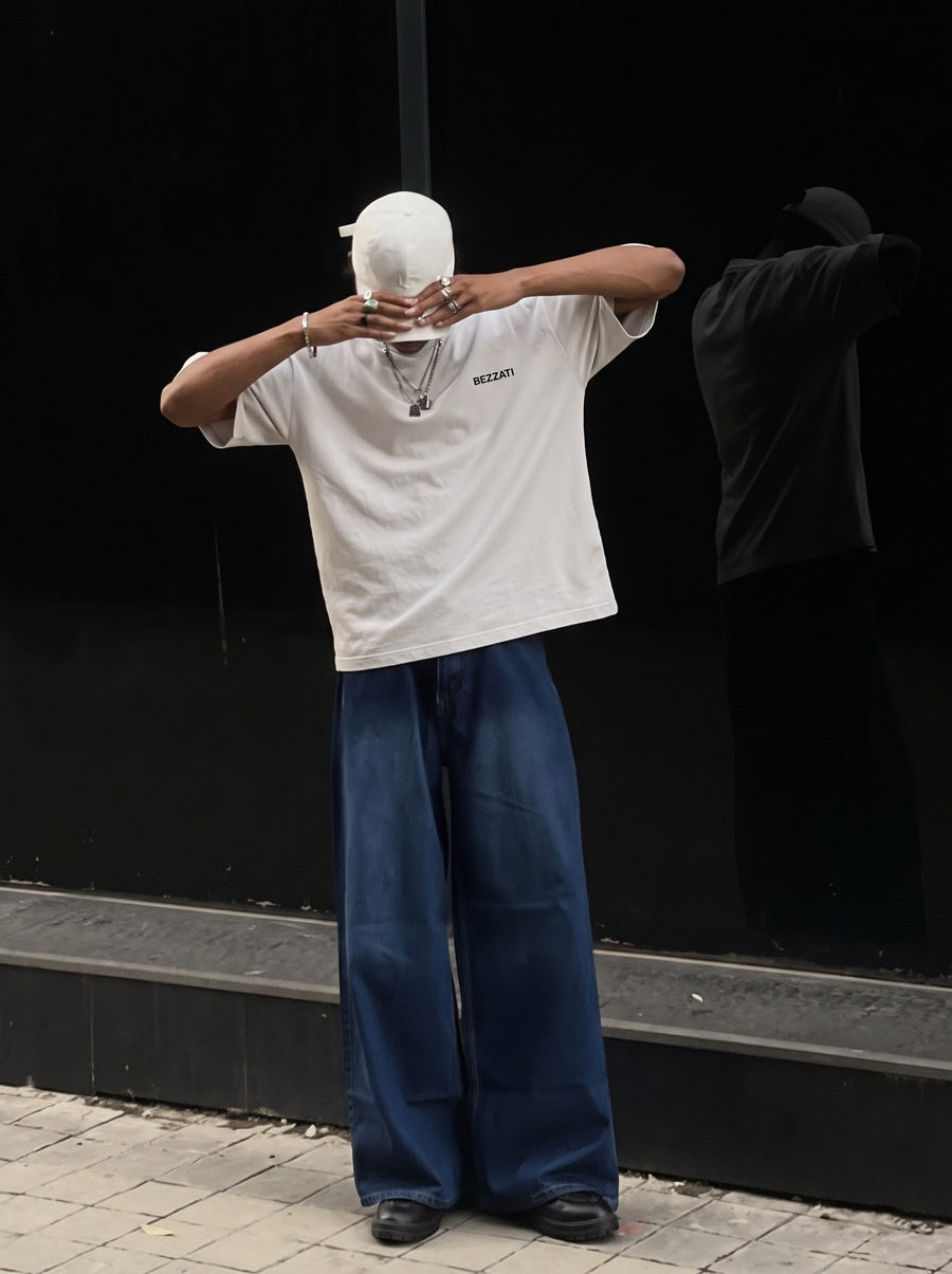 An off-white oversized tee worn with wide jeans against a dark wall, arms raised.